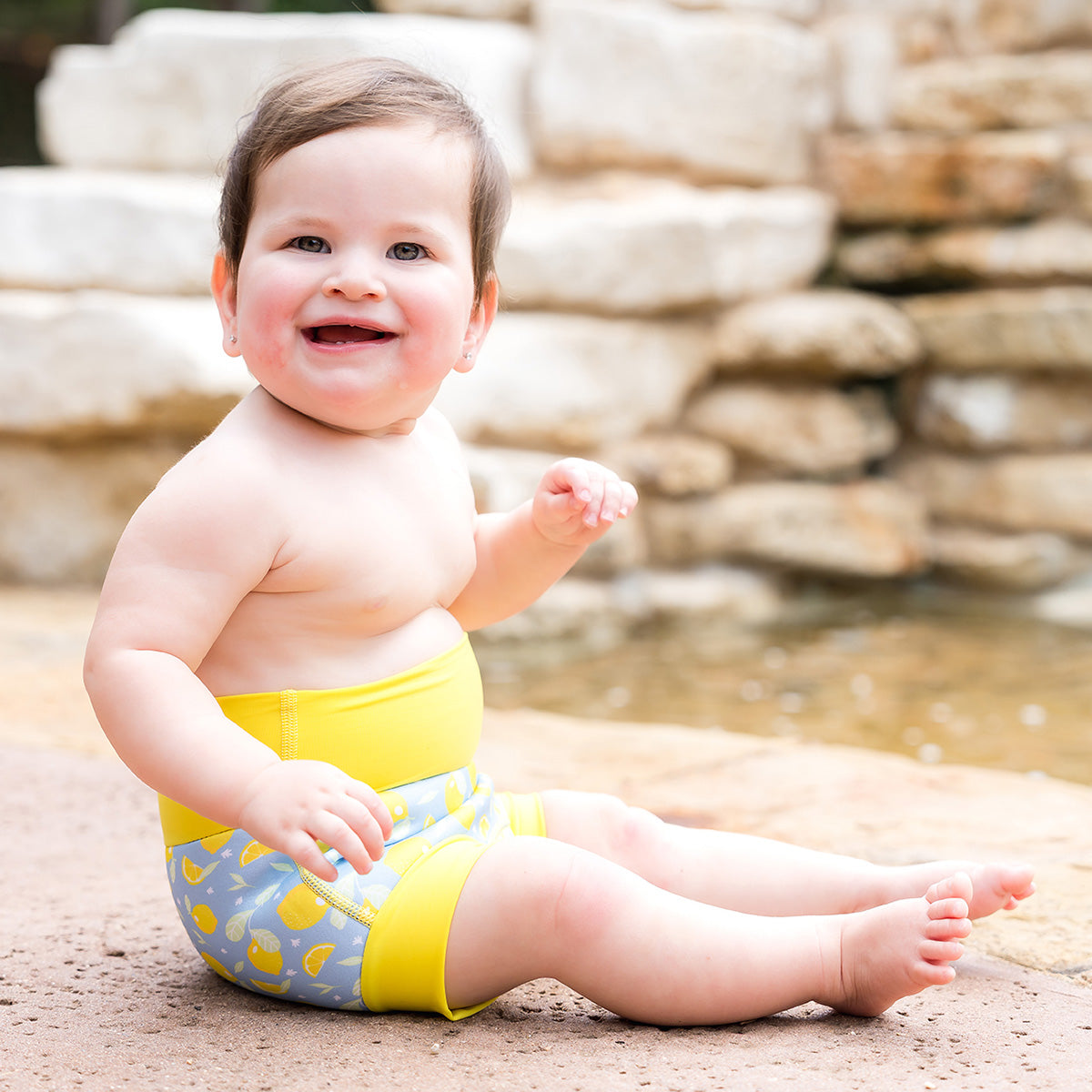 baby sat near the water wearing a lemon print happy nappy swim diaper