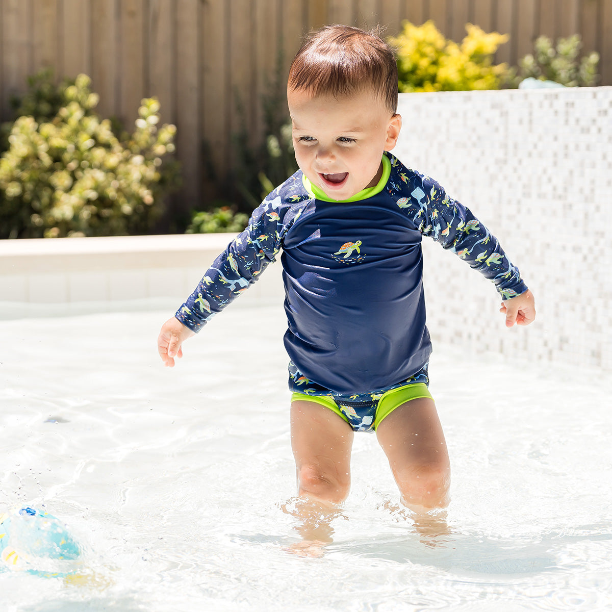 little boy wearing the turtle tide long sleeve rash top with matching happy nappy stood in the pool 