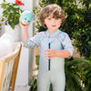 little boy stood infront of green garden holding a blue pufferfish toy and wearing a playful otter shorty wetsuit 