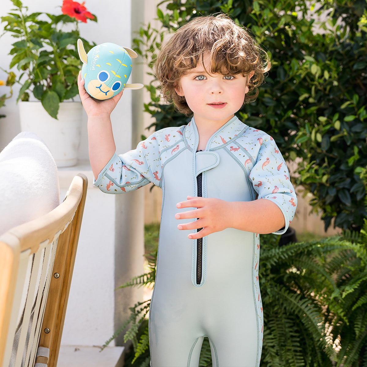 little boy stood infront of green garden holding a blue pufferfish toy and wearing a playful otter shorty wetsuit 