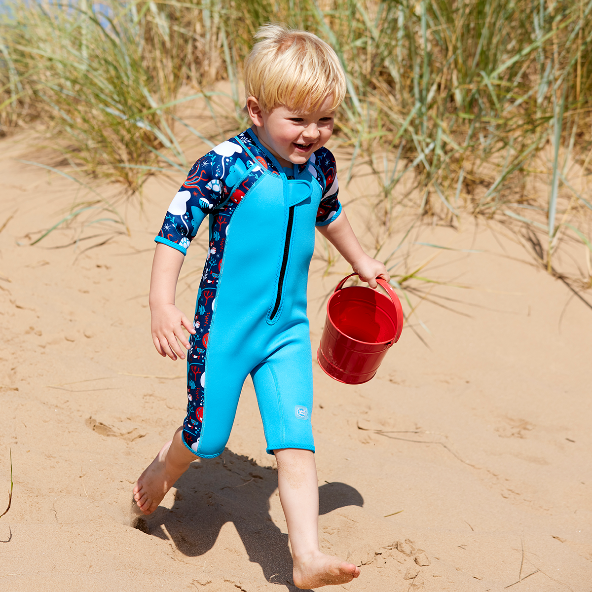 little boy running on the beach carrying a bucket wearing the shorty wetsuit under the sea
